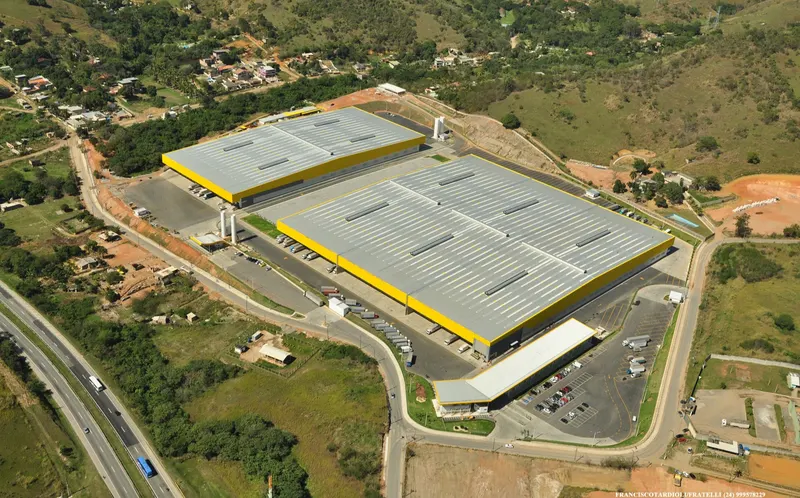 Vista aérea do CD Casa e Vídeo em Queimados/RJ: dois galpões com cobertura metálica, docas, pátio de caminhões e estacionamento.